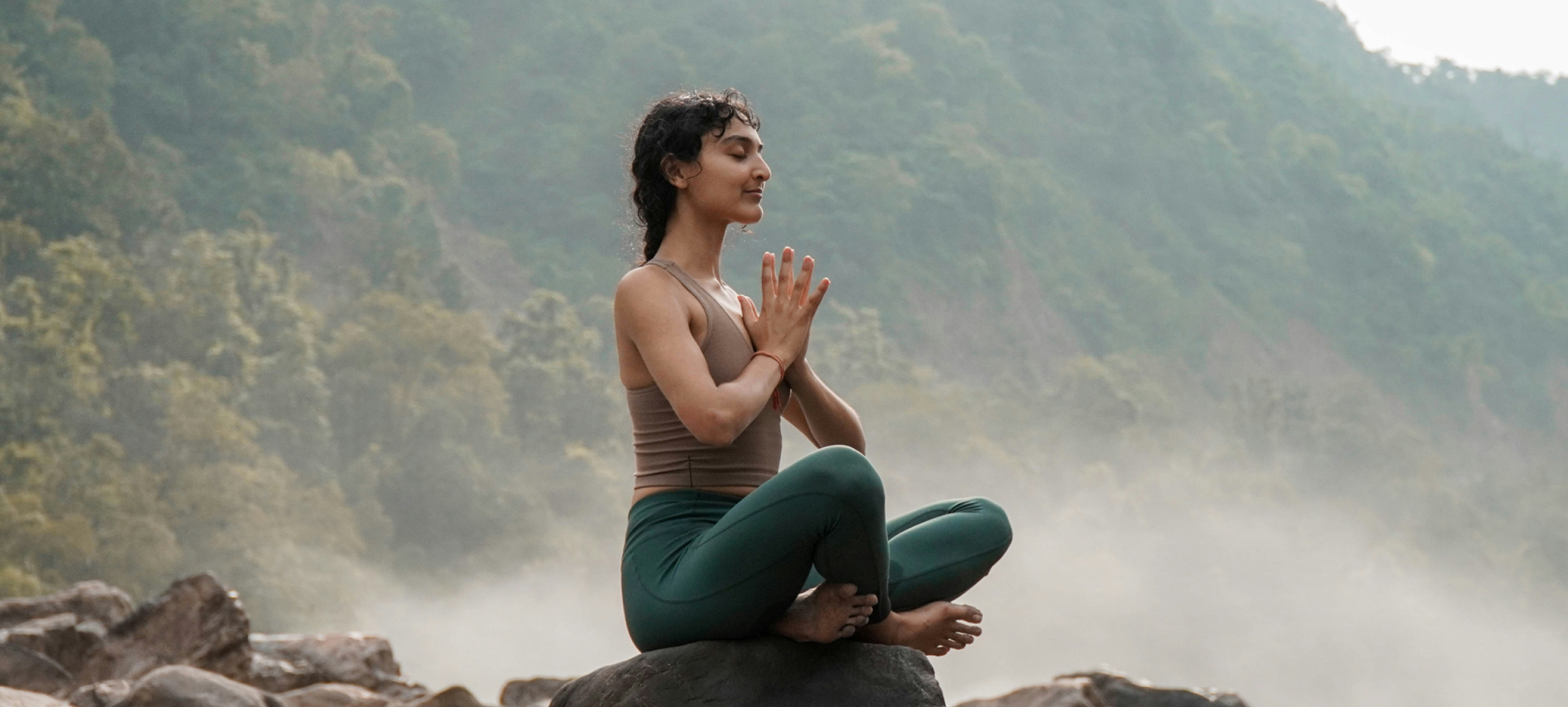 A woman sitting cross-legged in a peaceful outdoor setting, meditating with eyes closed and hands resting on her knees