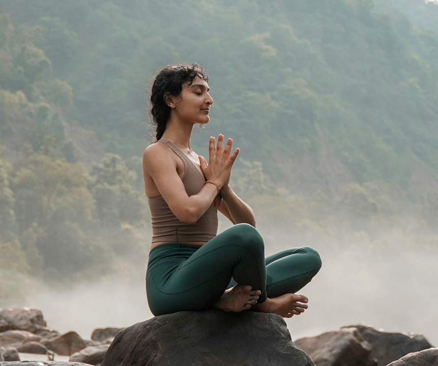 A woman sitting cross-legged in a peaceful outdoor setting, meditating with eyes closed and hands resting on her knees